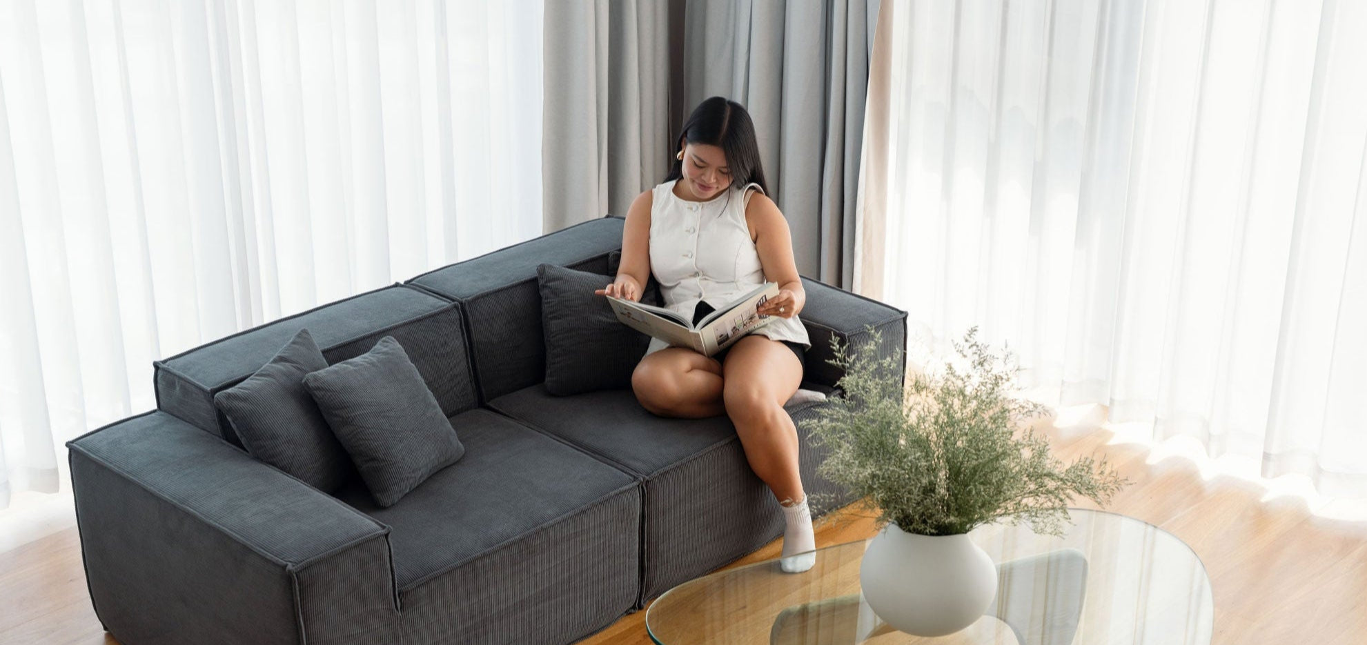Woman sitting on a gray sectional sofa reading a book in a bright living room.